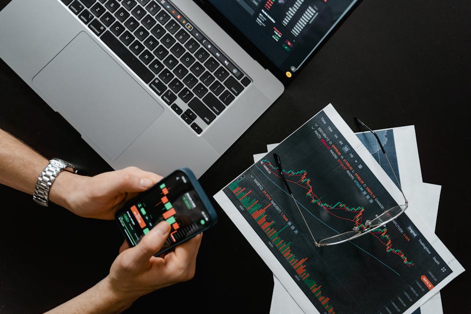 Hands using smartphone beside laptop with stock charts, showcasing digital trading